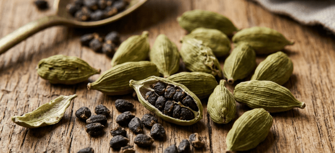 Green cardamom pods with some opened showing black seeds scattered on a wooden surface